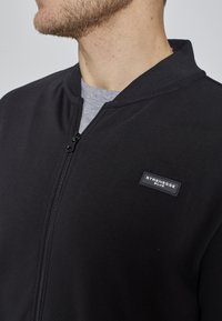 Veste noire à fermeture éclair, confectionnée dans un matériau lisse, avec un col côtelé et un patch logo Strenesse Blue sur le côté gauche.