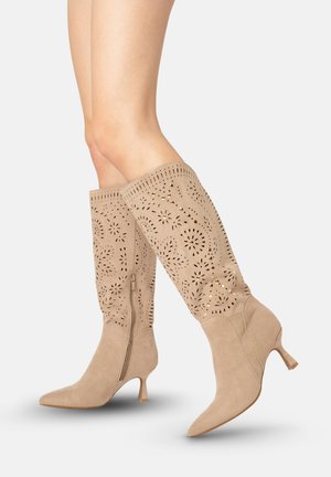 Ben iført beige ruskind støvler til knæet med laserudskårede blomsterprint, gyldne nitter, sidelukning med lynlås og lave hæle på en ensfarvet baggrund.