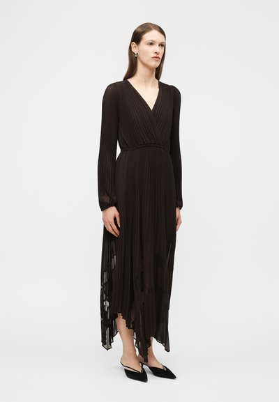 Γυναίκα με μακριά ίσια μαλλιά που φοράει μαύρο, μακρυμάνικο, πλισέ φόρεμα τύπου wrap με ασύμμετρο τελείωμα και μαύρες μυτερές μπαλαρίνες.