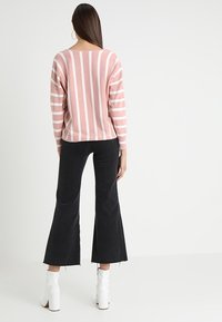 Lichtroze gestreepte trui met lange mouwen, gedragen over zwarte flared jeans. Schoenen zijn witte enkellaarzen met hak.