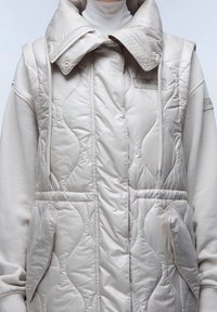 Světlé béžové prošívané vesta s vysokým límcem, předními kapsami a nastavitelnými šňůrkami. Vyrobená z lesklé, hladké látky s texturovaným povrchem.