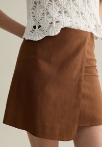 Brauner Wickelrock aus strukturiertem Stoff, mit geradem Schnitt und asymmetrischem Saum. Das Design ist minimalistisch und funktional.