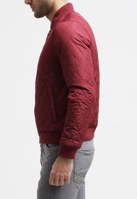 Quilted maroon bomber jacket with ribbed collar and cuffs, featuring side pockets and smooth texture. Simple design with functional detailing.