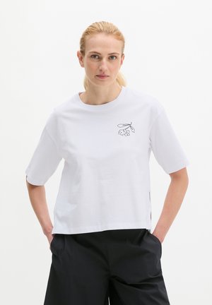 Kobieta o blond włosach ubrana w białą koszulę z krótkim rękawem z małym czarnym kwiatowym wzorem na piersi oraz czarne spodnie, ręce w kieszeniach.