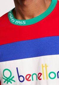 Camiseta bloqueada en colores rojo, azul y blanco con un cuello de canalé verde, que presenta el logo "benetton" y el texto 1965 en colores variados. Material de algodón suave.