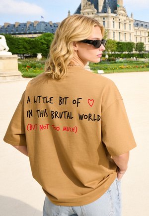 Blond persoon met zwarte zonnebril en beige shirt met tekst, staand buiten bij historische gebouwen en groene tuin onder een heldere lucht.