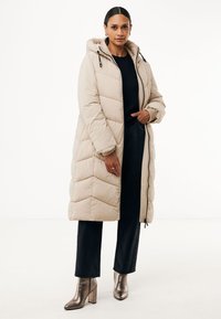 Beige pufferjas met capuchon, gewatteerde textuur, voorzijde met rits en trekkoorden. Gepaard met zwarte broeken en metalen haklaarzen.