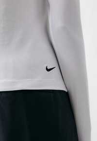 Nike garā piedurkņu krekls ar gludu gaiši pelēku audumu, melniem šuvēm un mazu melnu logotipu apakšmalā aizmugurē.