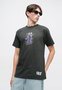 Młody mężczyzna w ciemnych okularach przeciwsłonecznych, czarnej koszulce z grafiką "The Nightmare Before Christmas" i jasnoniebieskich dżinsach, patrzy w bok.