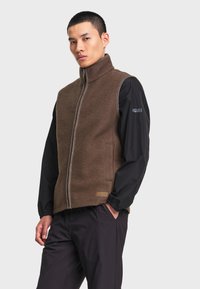 Homme portant un gilet polaire marron zippé sur une veste noire à manches longues, debout avec une main dans la poche devant un fond uni.