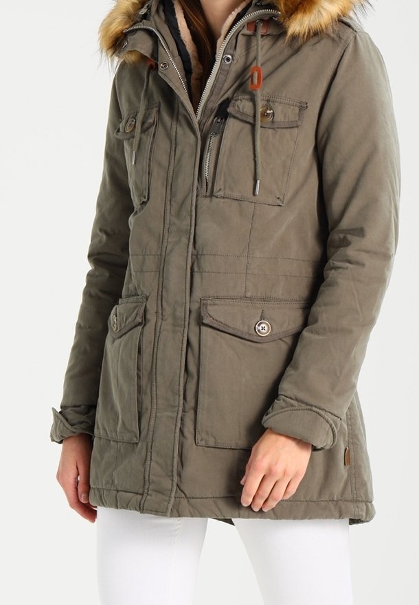Femme portant une parka vert olive avec une capuche bordée de fourrure, plusieurs poches avant et une fermeture à cordon, associée à un pantalon blanc.