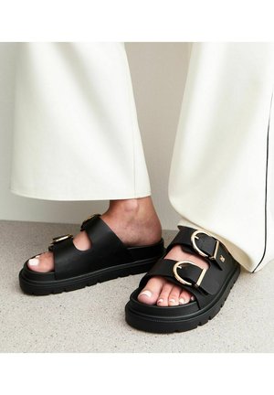 Svarta sandaler med två breda remmar som har runda guldspännen. Tjock sula med en texturerad yta. Tårna är exponerade och har en ljus hudton.