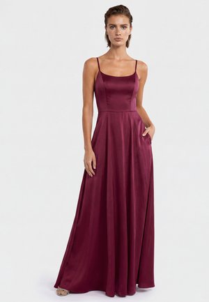 Woman wearing a sleeveless, floor-length burgundy satin dress with thin straps, standing straight with one hand in the dress pocket.