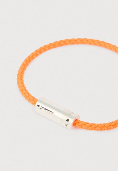 Oranžno pleteno zapestnico z valjastim srebrnim zapiranjem, na katerem je vgravirano "le gramme" in oznake teže, na beli podlagi.