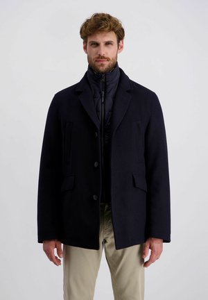 Homme barbu aux cheveux bouclés portant un blazer noir superposé sur une veste matelassée zippée et un pantalon beige, sur un fond uni.
