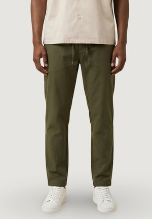 Uomo che indossa pantaloni con coulisse verde oliva, camicia beige e sneakers bianche, in piedi davanti a uno sfondo semplice.
