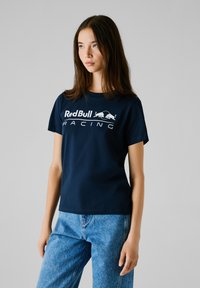 T-shirt in cotone blu navy con logo "Red Bull Racing" in bianco, maniche corte e design classico con scollo a girocollo, abbinata a jeans di denim blu chiaro.