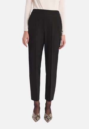 Kobieta w czarnych spodniacach typu tapered, cienkich czarnych rajstopach, butach na obcasie w wężowy wzór i białej bluzce z długim rękawem, stojąca na tle białym.