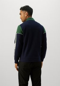 Sweatshirt azul-marinho com detalhes verdes e colarinho alto. Apresenta rebordos brancos e detalhes em padrão nas mangas. Textura suave.
