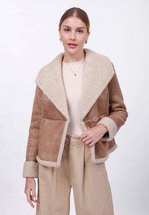 Beige schapenvachtjack met een crèmekleurige neef van nepbont, bijpassende manchetten en twee knopen. Geribbeld lichtbeige top en beige broeken zijn zichtbaar.
