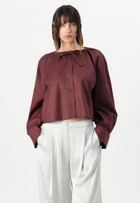Burgundy cropped shirt with a neck tie, button closure, and wide sleeves, paired with light gray high-waisted trousers. Simple design.