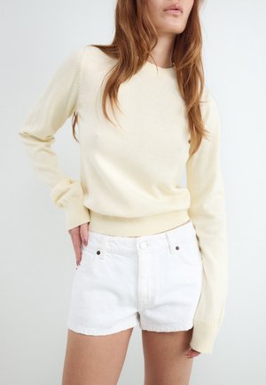 Femme portant un pull crème à manches longues et un short en denim blanc, la main gauche posée sur la hanche.