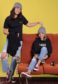 Due bambini indossano berrettini grigi, una maglietta nera e una felpa nera con un logo. Sono seduti su una sedia a strisce arancioni con dei jeans e sneakers rosse.
