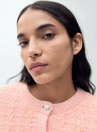 Svetlo roza teksturirana jakna z okroglim ovratnikom, ki ima srebrno gumbno zapiranje in mehko tkano blago. Vidna gladka koža in temni lasje.