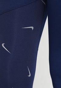 Leggings de sport bleu marine en tissu lisse et extensible, avec un logo Nike argenté et des motifs discrets de swoosh sur toute la surface.