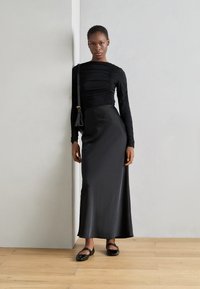 Schwarzes langärmeliges Kleid mit gerafftem Detail am Oberteil, glattem Satinstoff und figurbetontem Schnitt. Schwarze flache Schuhe runden den Look ab.