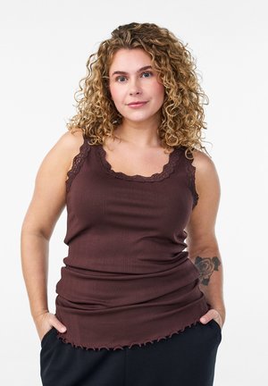 Vrouw met krullend blond haar, een mouwloze bruine top en zwarte broek, handen in de zakken, met een rozentatoeage op haar arm.