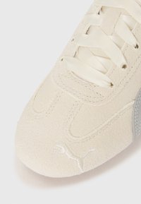 Sneaker bianco in suede con superficie testurizzata, caratterizzato da lacci beige chiaro e un accento in logo argentato. Punta arrotondata con dettagli di cucitura.
