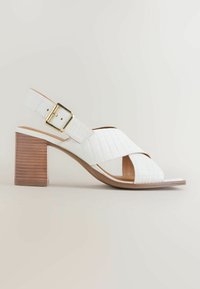 Sandale slingback en cuir blanc avec texture embossée, boucle dorée, brides croisées et talon bloc en bois empilé sur fond neutre.
