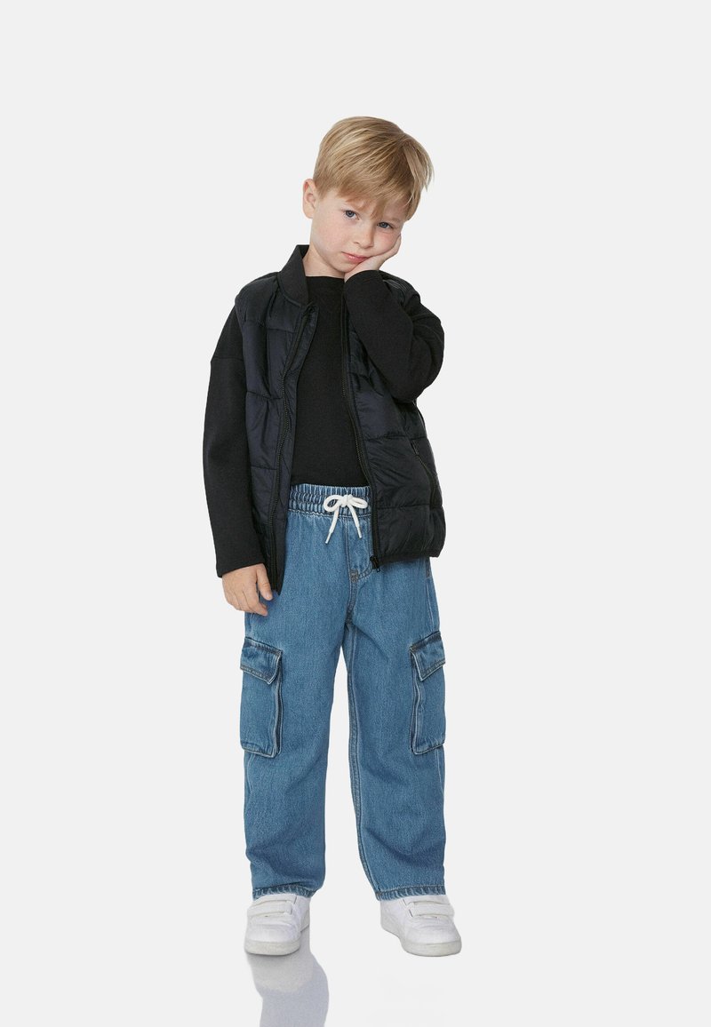 Dítě v černém puffer vestě přes černé dlouhé tričko, doplněné volnými světlými modrými cargo kalhotami a bílými teniskami.