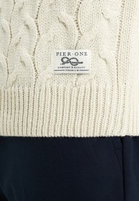 Maglione crema a maglia cable con orlo a coste e un'etichetta di tessuto con la scritta "PIER ONE". Abbinato a pantaloni blu scuro, mostrando colori a contrasto.