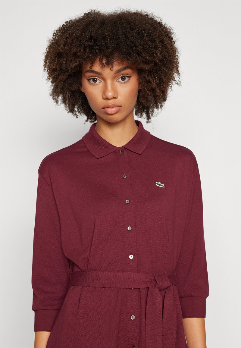 Burgunderfarbendes Hemdkleid mit Kragen, Dreiviertelärmel, gebundenem Bund und kleinem grünen Logo. Texturiert mit einer figurbetonten Passform.