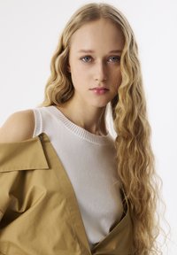 Beżowa, oversize'owa kurtka o matowym wykończeniu nałożona na białą, prążkowaną bluzkę z dzianiny. Modelka ma długie, falowane blond włosy na tle neutralnym.