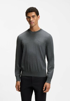 Giovane uomo che indossa un maglione aderente a maniche lunghe grigio scuro e pantaloni neri, in piedi davanti a uno sfondo bianco semplice.