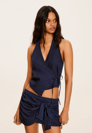 Kvinde med langt mørkt hår iført navyblå ærmeløs omslags crop top og matchende bundet mininederdel, stående mod en lys baggrund.