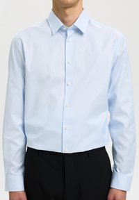 Hombre con camisa azul claro de manga larga abotonada metida en pantalones negros, sobre un fondo liso.