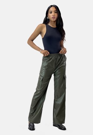 Schwarzer ärmelloser Bodysuit, olivgrüne Fake-Leder Cargo-Hosen mit weitem Bein und seitlichen Taschen, schwarze verzierten flache Schuhe, minimaler Schmuck.