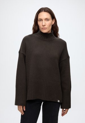 Femme aux longs cheveux portant un pull à col roulé côtelé marron foncé et un pantalon noir, debout devant un fond clair uni.
