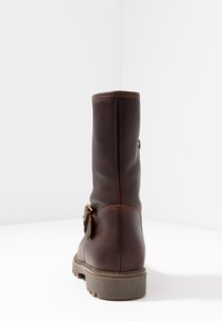 Botte unique en cuir marron foncé montant mi-mollet avec une boucle sur le côté et une semelle épaisse en caoutchouc, photographiée de dos sur une surface blanche.