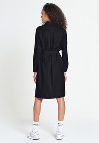 Abito camicia nero con cintura, maniche lunghe, colletto e taglio dritto. Tessuto liscio e design fino al ginocchio. Sneakers bianche e calze visibili.