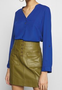 Blå blus med V-hals och knapppdetaljer, som matchas med en hög midja olivgrön läderkjol med framfickor och dekorativa knappar.