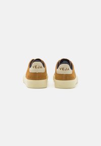 Brune semskede sneakers med hvite gummisåler, med en marineblå tekstilkrage og "VEJA"-logoet bak i brune og kremfargede toner.