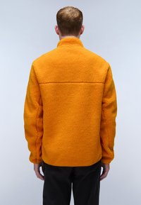 Orange fleece jacket with a high collar, textured fabric, and seaming details. Designed with long sleeves and a relaxed fit.