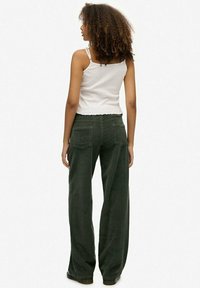 Pantalones de pana de pierna ancha en verde oscuro, con dos bolsillos traseros, tejido texturizado y cintura alta. Combinados con una blusa blanca ajustada.