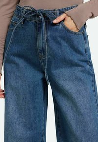 Pantalones de mezclilla de pierna ancha con cinturón de lazo, en un color azul medio, con un leve desgastado y bolsillos frontales. Tela de algodón texturizada.