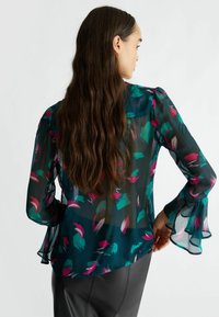 Blusa trasparente di un verde bluastro scuro con una stampa di piume colorate in rosa e verde. Presenta maniche svasate e una silhouette morbida. Tessuto texturizzato.
